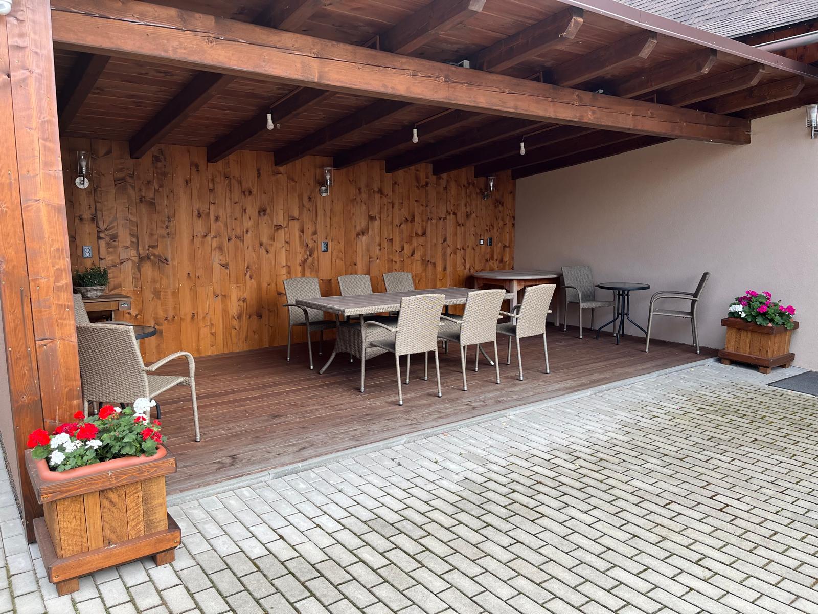 Krytá terasa obložená dřevem s dlouhým stolem, křesílky a truhlíky s muškáty