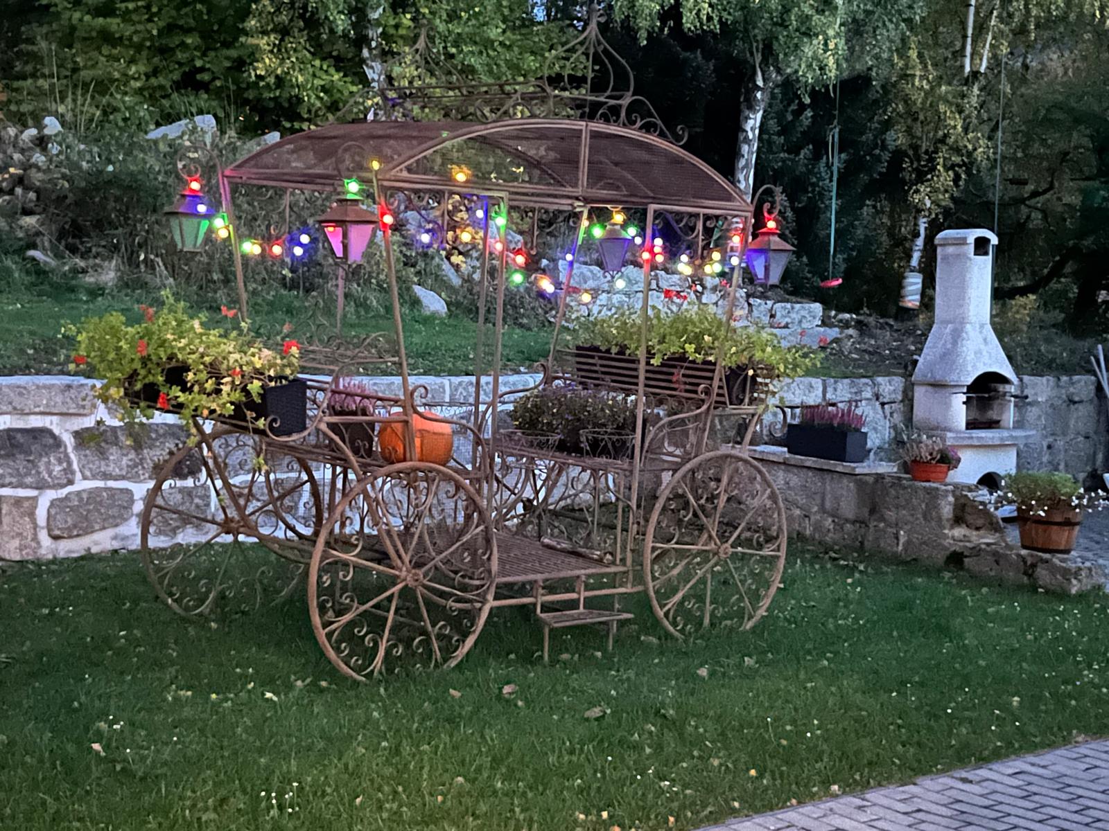 Kovový kočár s květinami a barevnými světýlky na zahradě za soumraku
